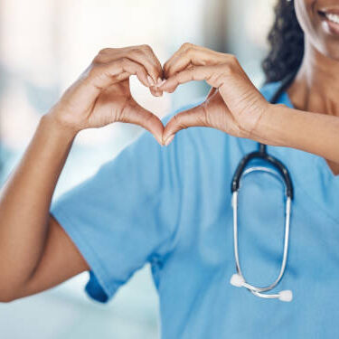 istockphoto-1400388878-612x612 Closeup african american woman nurse making a heart shape with her hands while smiling and standing in hospital. Take care of your heart and love your body. Health and safety in the field of medicine